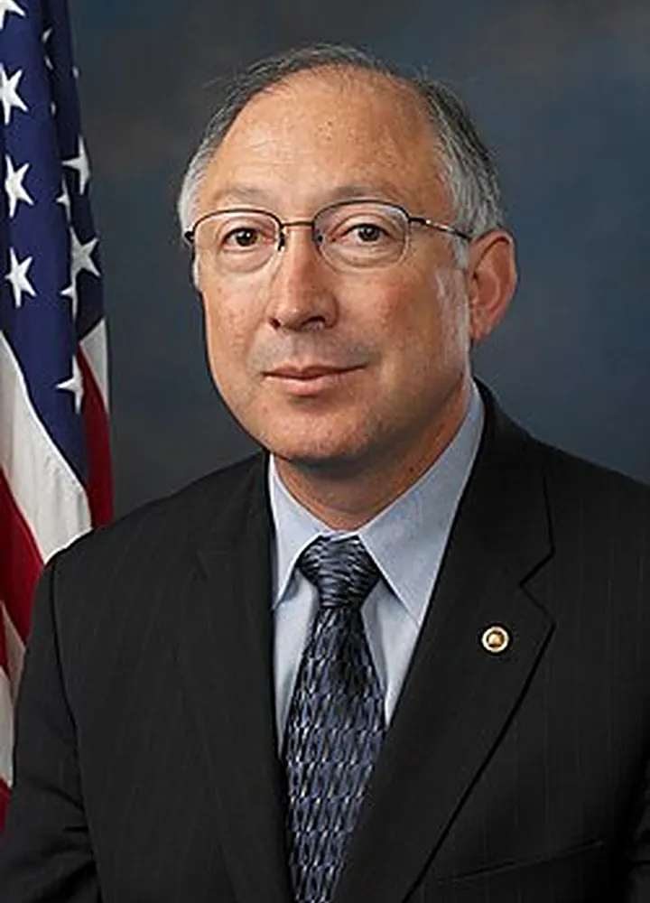 Official portrait of Ken Salazar in a dark suit in front of an American flag