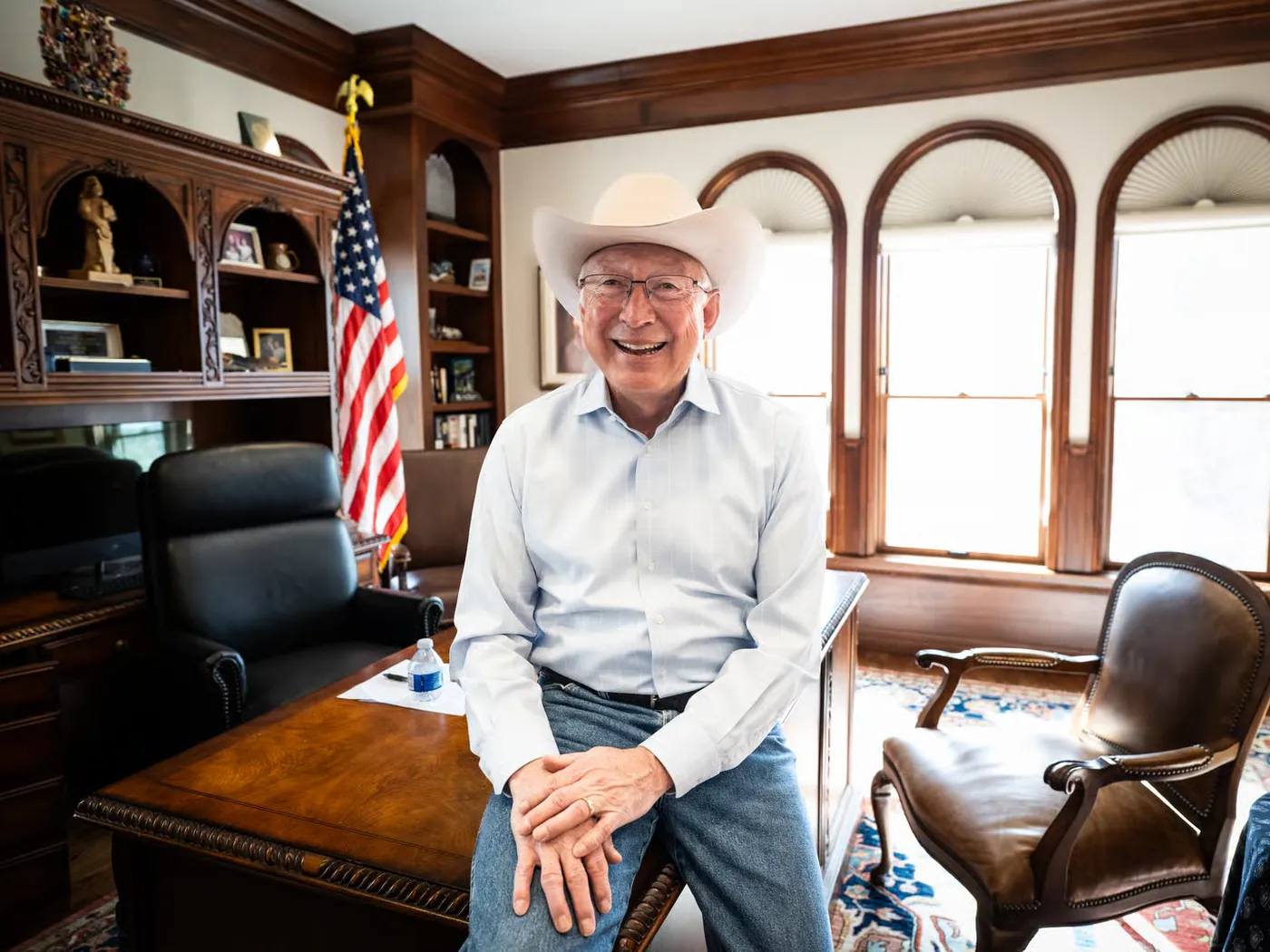 Ken Salazar laughing in his cowboy hat, seated on the edge of the desk in his office with an American flag behind him